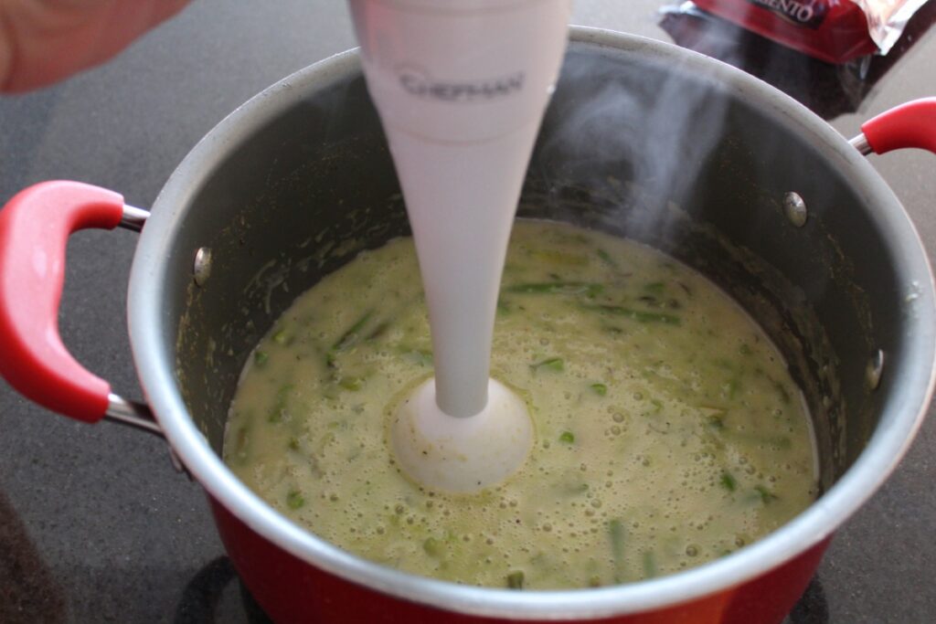 a pot with asparagus soup in it with an immersion blender placed in the soup in a pot o top of a counter.