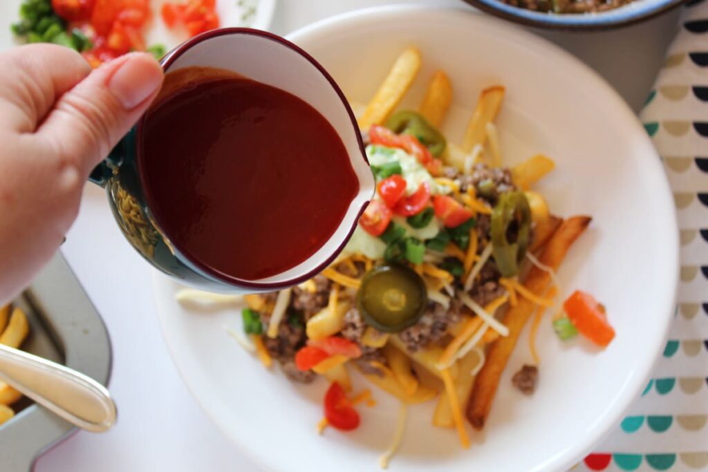 cup pouring taco sauce over the top of french fries loaded with ground beef and taco toppings.
