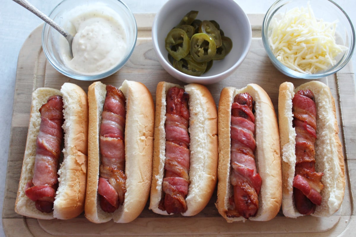 Two hot dogs with bacon wrapped around them and toppings on top.