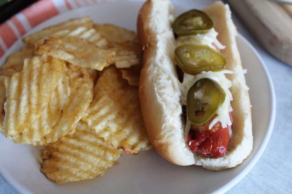 all beef hot dogs wrapped in bacon in hot dog buns on a plate with potato chips next to them.