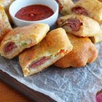 homemade Stromboli made with pizza crust and is sliced and placed on a cutting board and a piece of parchment paper. a bowl of pizza sauce in next to it.