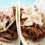 side zoomed in shot of beer-braised pork tacos with apple coleslaw on top in soft tortillas.