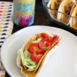 a plate with a taco on it with a pan of double deluxe tacos a beer and a cloth napkin next to it.