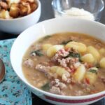 a bowl of gnocchi sausage and spinach soup wiht a cloth napkin and spoon next to it and bowls of croutons and parmesan cheese in the background