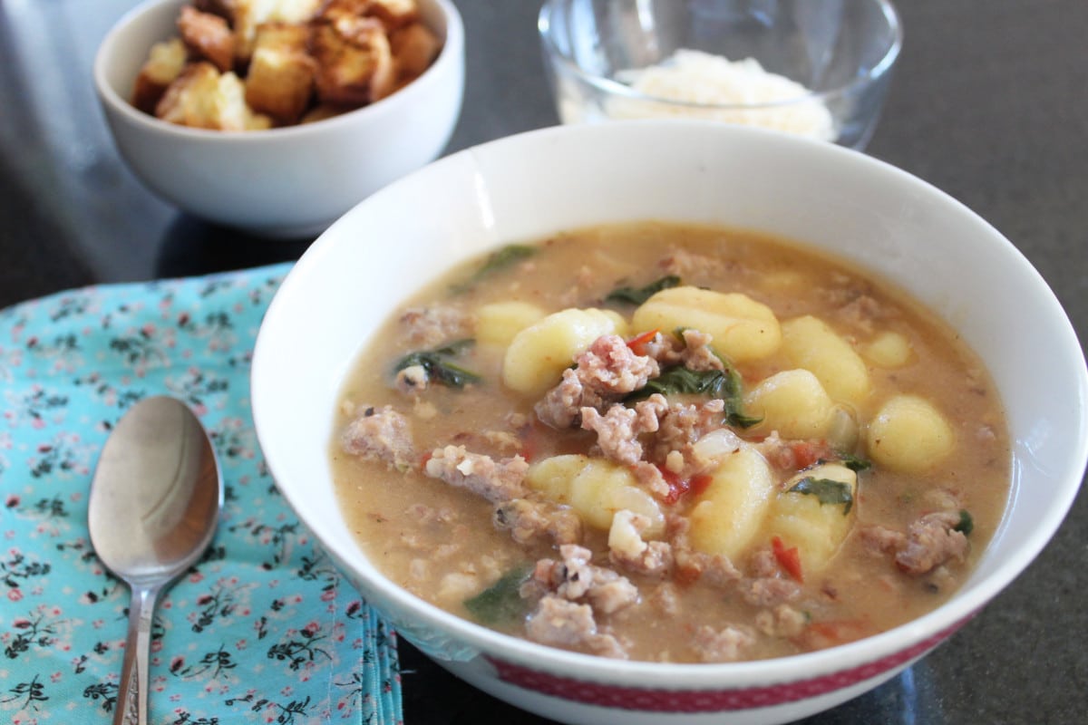 long shot of a bowl of gnocchi sausage soup with parmesan and croutons and a cloth napkin next to it