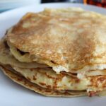 Swedish Pancakes stacked on a plate with a jar of maple syrup behind it.