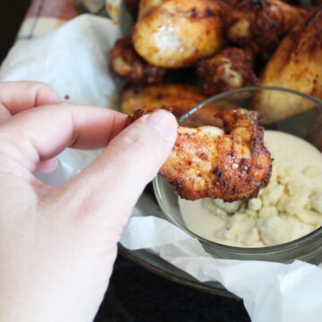 zoomed in shot of a hand dipping a chicken drummy into a a dish of blue cheese dressing. more chicken wings in the basket and c loth napkin next to it.