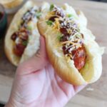 a hand holding a bacon wrapped hot dog covered with BBQ sauce and cheese over a board holding more hot dogs and toppings.