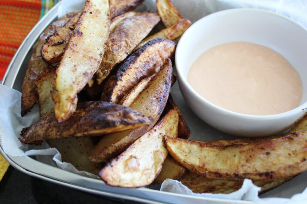 side shot of air fryer spicy potato wedges in a container with wax paper and a bowl of French fry dipping sauce in a bowl.