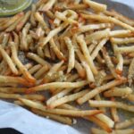 zoomed in side shot of french fries drizzled with pesto sauce mixed with butter and parmesan cheese on a piece of parchment paper on a pan.